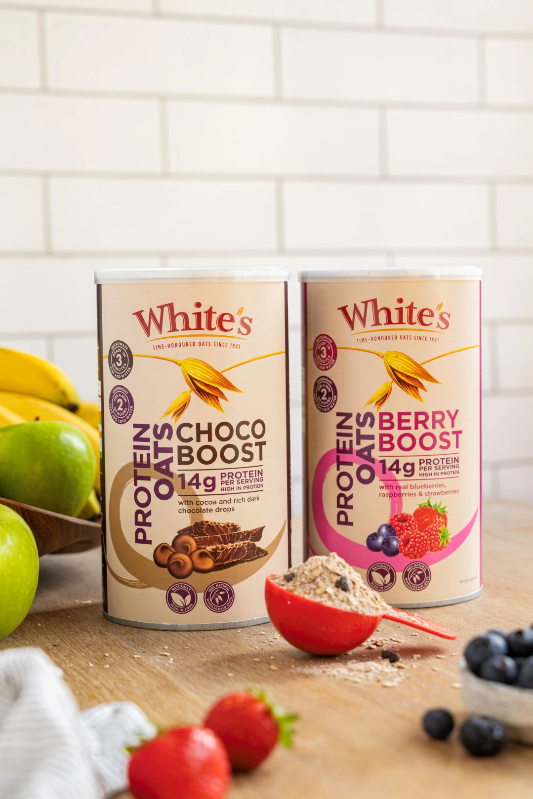 Two packages of White's new protein oats product sat on a wooden surface against a white tiled background. There is a spoonful of oats and mixed berries in the foreground.